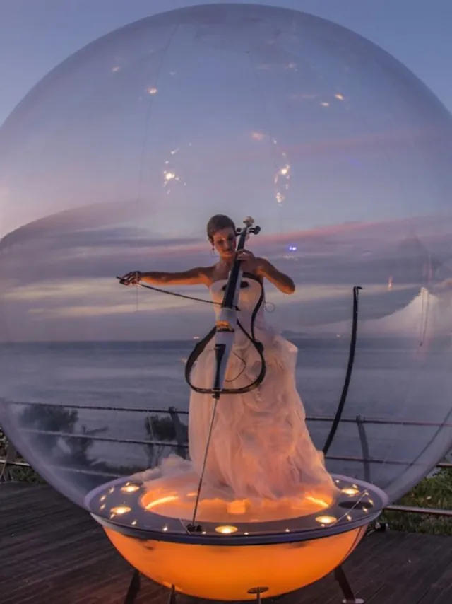 Cellist in a white gown inside a lit bubble on a terrace above the sea